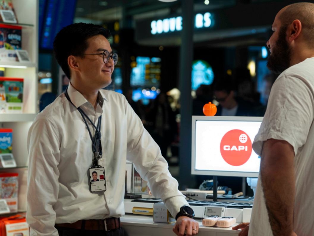 Butikkmedarbeidar møter ein kunde ved skranken i ein elektronikkbutikk, med produktutstilling og ein dataskjerm med Capi-logo i bakgrunnen