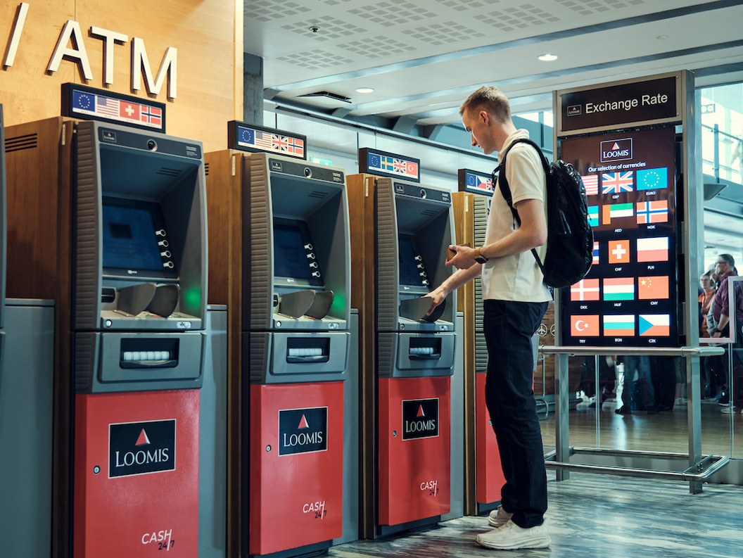 En person bruker en minibank for utenlandsk valuta på en flyplass, med flere valutaalternativer vist på en skjerm i nærheten, noe som indikerer internasjonale reiser og finansielle transaksjoner