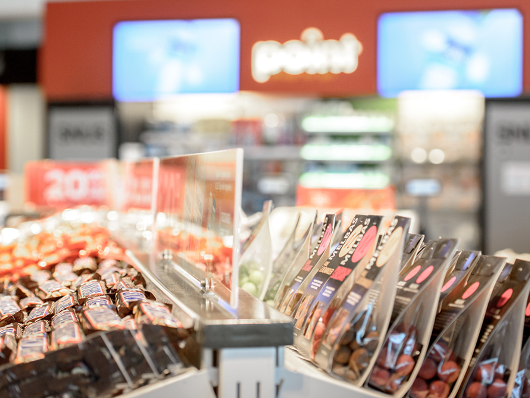 Snack display in a convenience store with various packaged candies and chocolates, featuring promotional signs and a beverage cooler in the blurred background