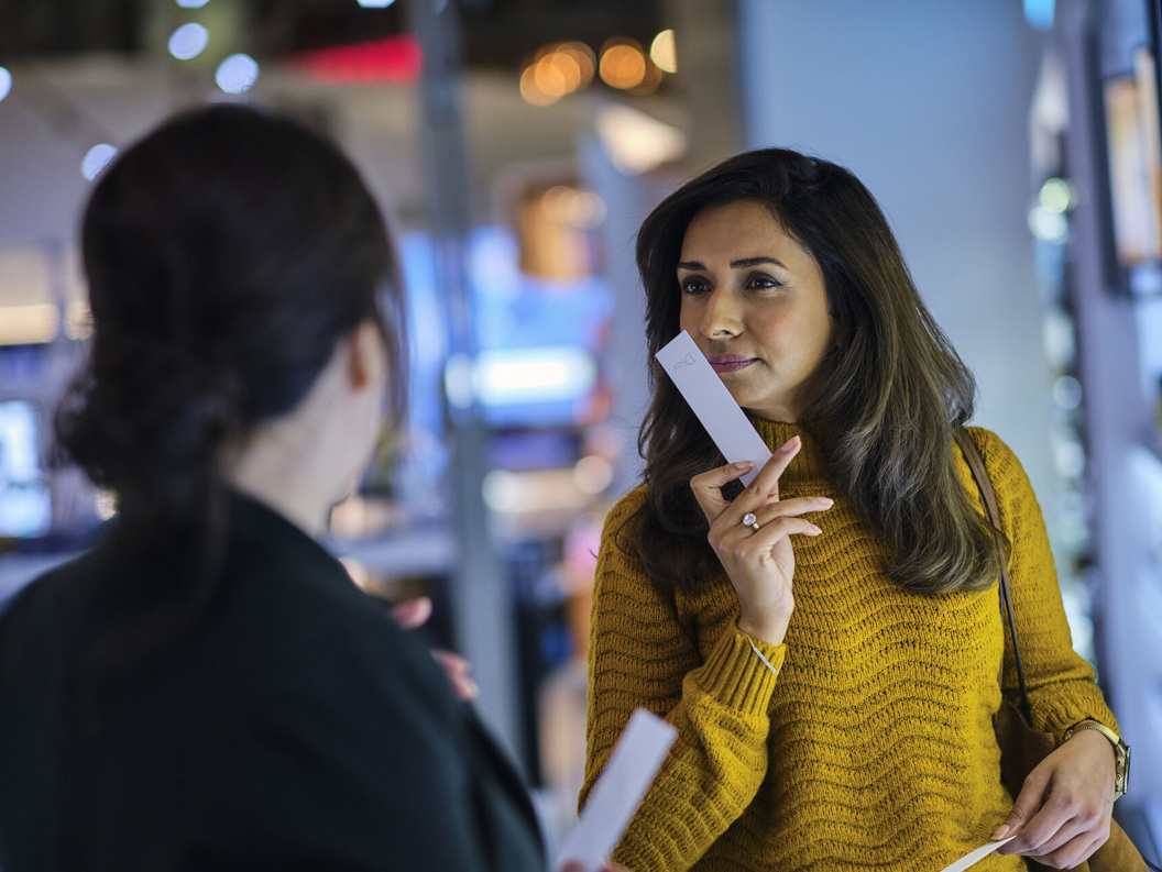 Woman in yellow sweater sampling perfumes in a store, holding a scent strip, with another person blurred in the foreground