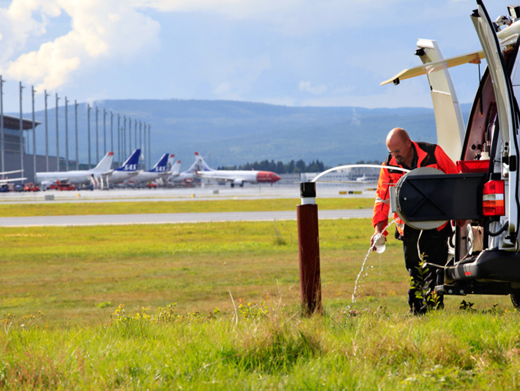 Mann i arbeidsklær fyller væske fra en hvit varebil ved en gressplen nær en flyplass, med flere fly på bakken i bakgrunnen.