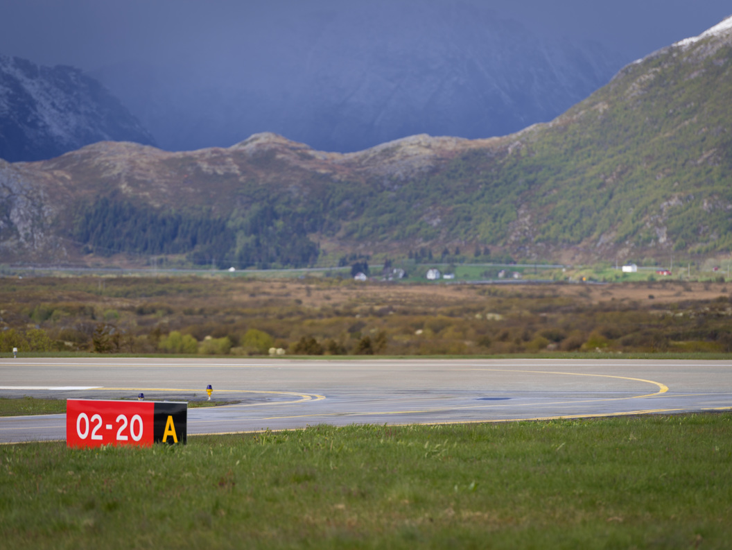 Flyplassrullebane med skilt 02-20 A, omgitt av norsk fjellandskap og grønne daler under overskyet himmel.