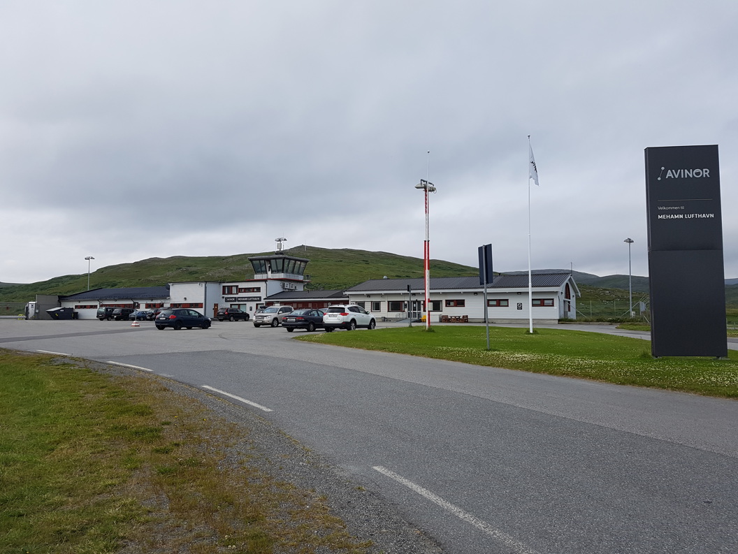 Mehamn lufthavn i Norge, med parkerte biler og kontrolltårn, omgitt av grønne åser under en overskyet himmel.