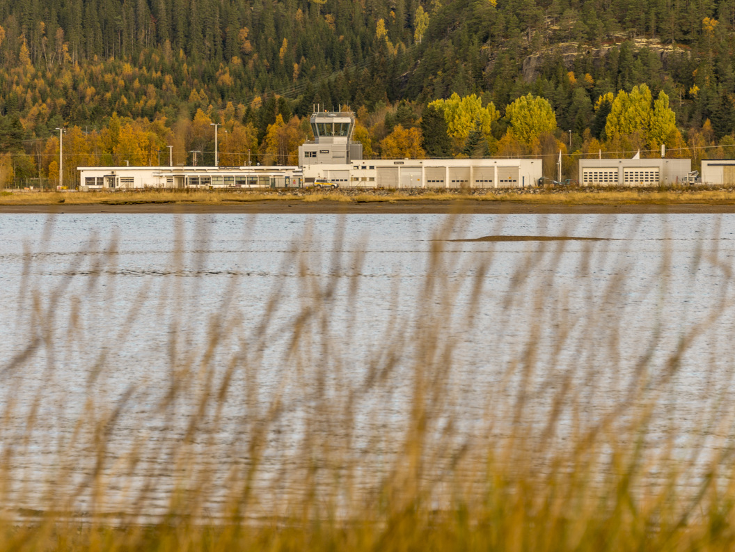 Flyplassbygning ved innsjø om høsten, omringet av høstfargede trær og åser i bakgrunnen, med siv i forgrunnen.