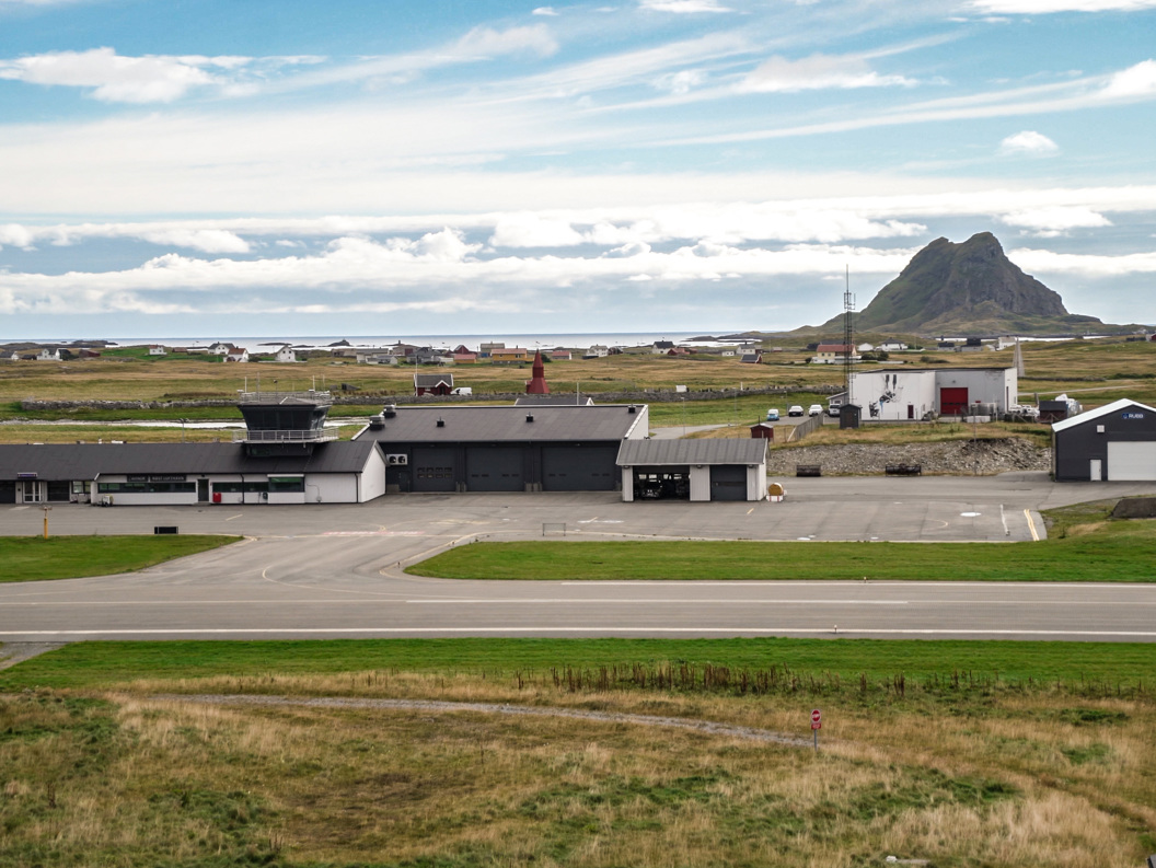 Flyplassbygning med landingsstripe i et landlig landskap med fjell i bakgrunnen under en delvis skyet himmel.