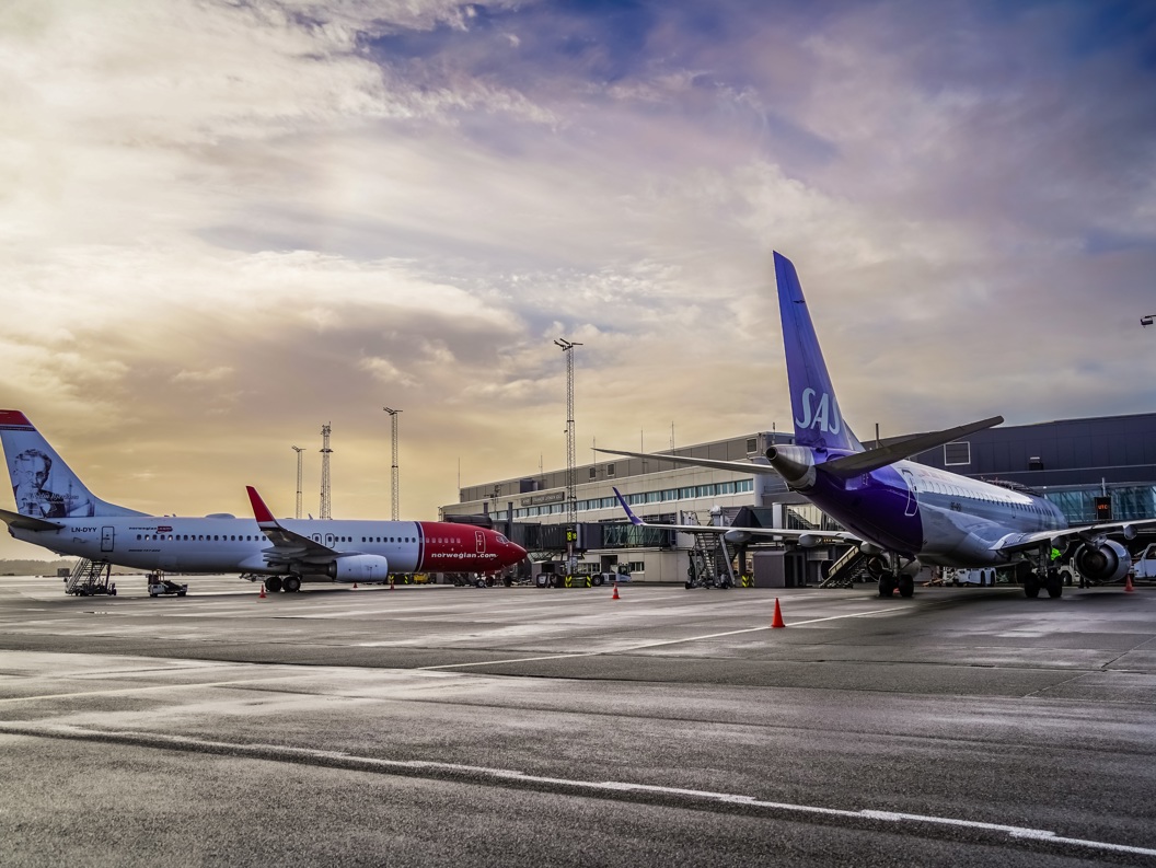 To fly på en flyplass i solnedgangen, et Norwegian-fly og et SAS-fly parkert ved en terminal, med et moderne terminalbygg i bakgrunnen.