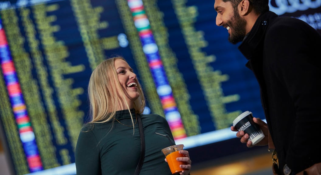 To personar smiler og held drikke i ein flyplass terminal framfor ein avgangstavel