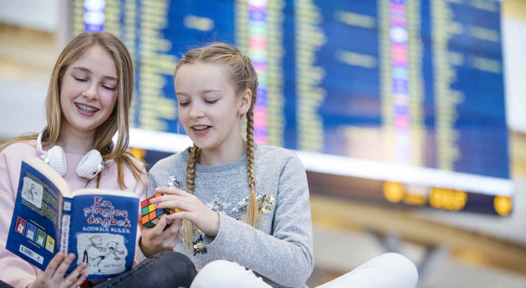 To unge jenter sit i ein flyplass med ein Rubiks kube og ei bok. Ei av jentene har hovudtelefonar rundt halsen. Bakgrunnen har ein stor informasjonstavle