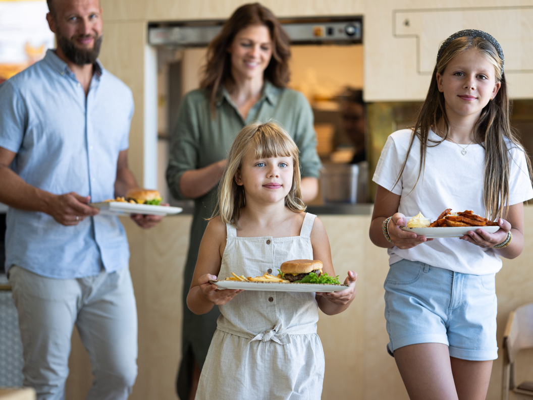 Familie på fire serveres måltider på en restaurant; far og mor i bakgrunnen, to døtre i forgrunnen med hamburgere og pommes frites på tallerkener.