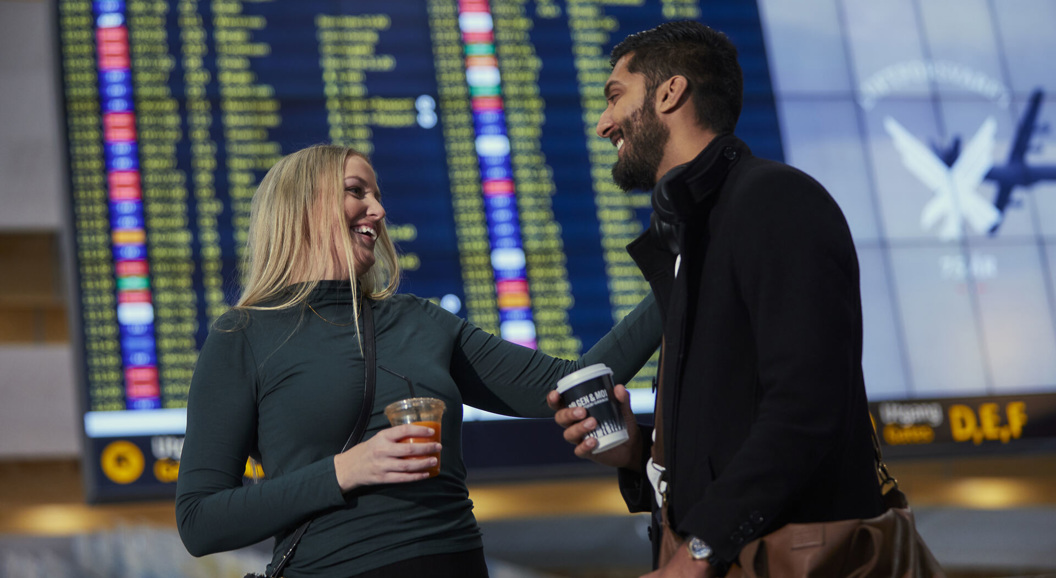 Two people at an airport are smiling and holding drinks, with a departure board visible in the background.
