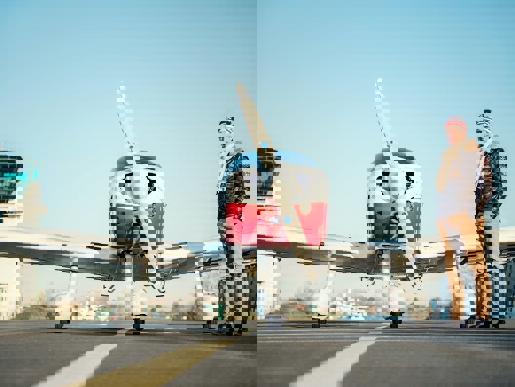 Lite propellfly på flyplass med kontrolltårn i bakgrunnen, stående mann kledd i vinterklær ved siden av flyet.