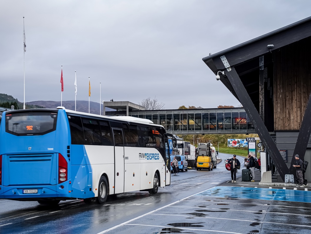 Blå buss på utenfor en flyplass med reisende, omringet av moderne arkitektur og fjellandskap i bakgrunnen.