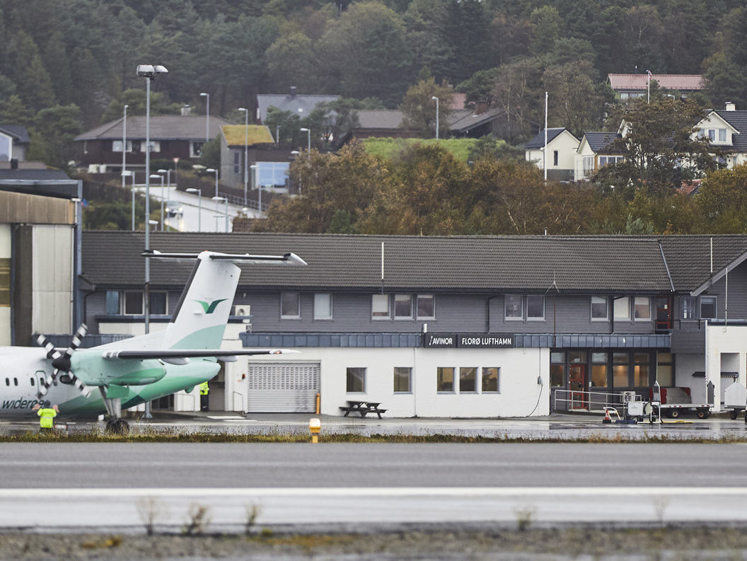 Widerøe-fly ved Florø lufthamn framfor terminalbygningen, omgitt av skog og bustader i bakgrunnen.