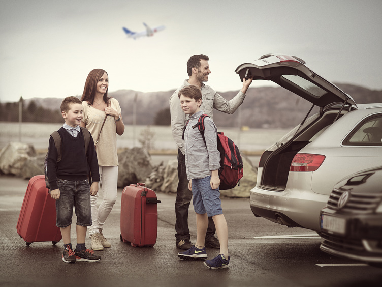 Familie på flyplass parkeringsplass med kofferter, bil og fly i bakgrunnen klar for reise.