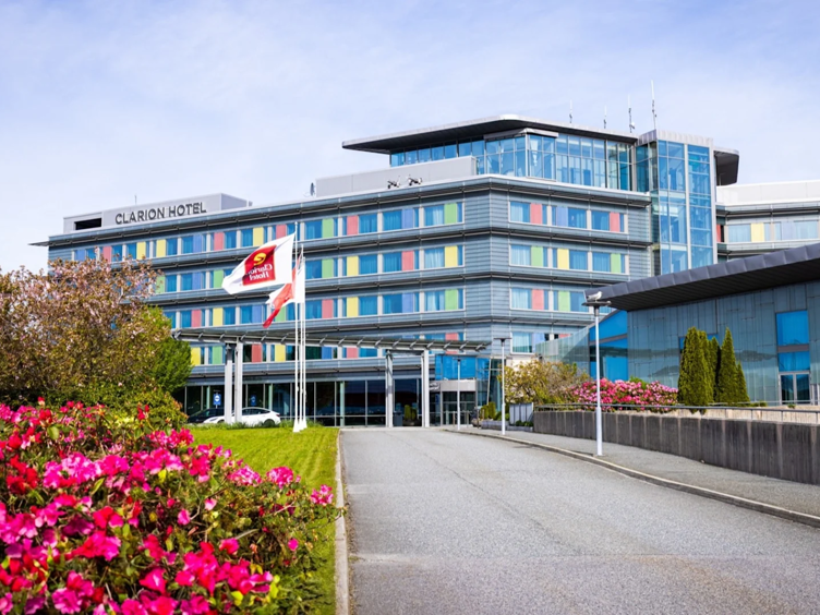 Moderne fasade av Clarion Hotel omgitt av frodige blomster og trær under en klar himmel.