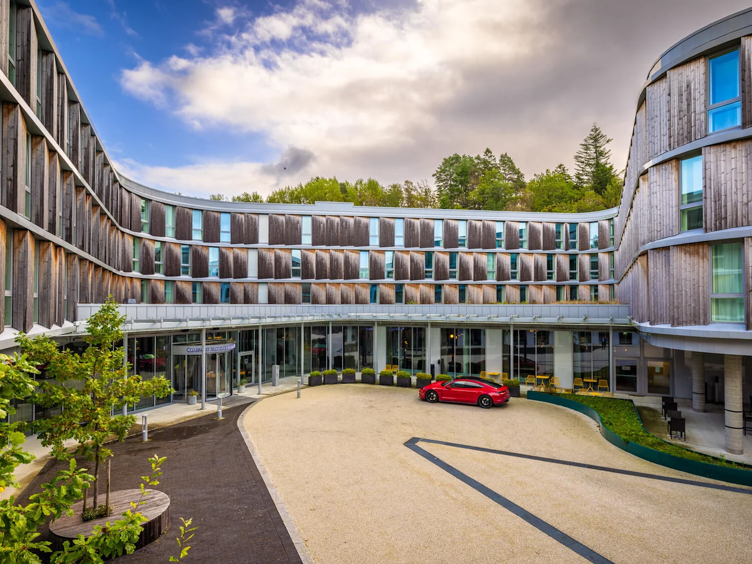 Moderne hotellfasade med buet design, store vinduer, og en rød bil parkert foran hovedinngangen, omgitt av grøntområder og delvis overskyet himmel.