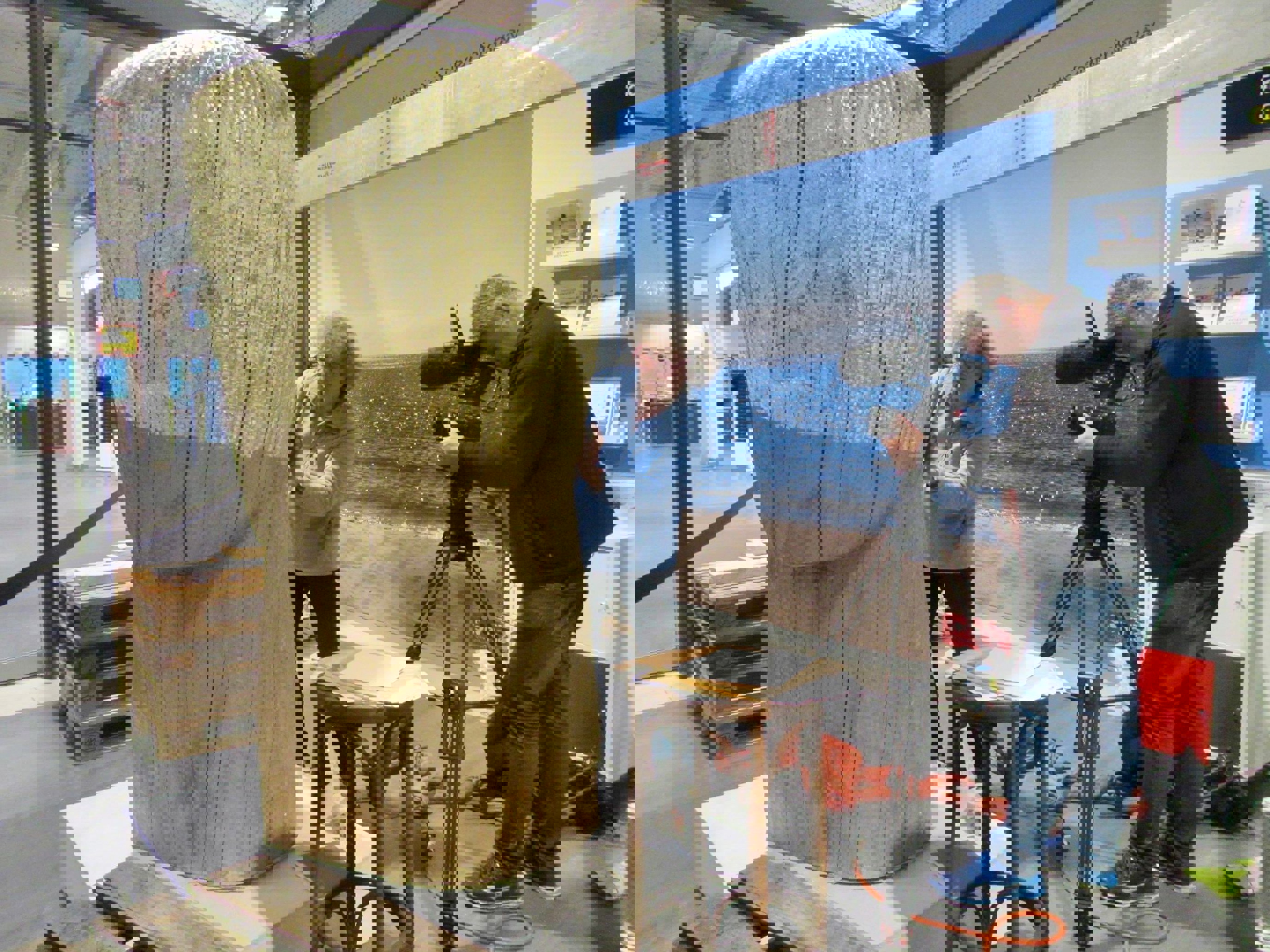 Kunstner arbeider på en stor treutskjæring mens en kameramann filmer. Scenen er innendørs med en bakgrunn av en strandpromenade fra Sørlige Norge