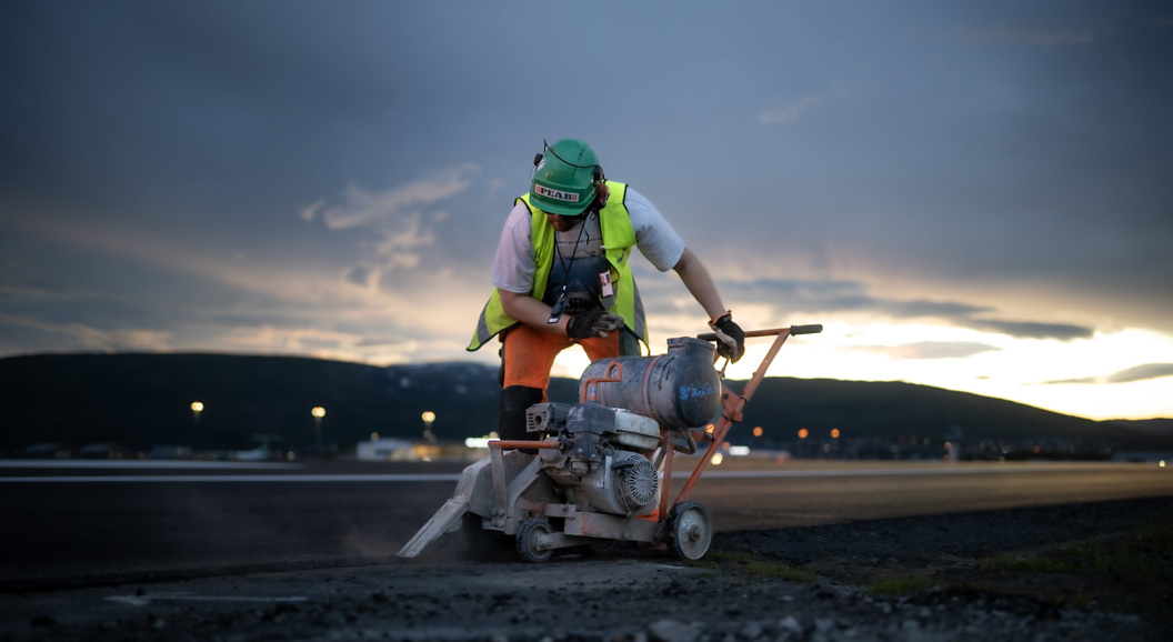 Arbeider kutter asfalt med betongsag på en byggeplass ved solnedgang.