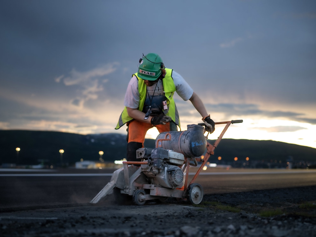 Arbeider kutter asfalt med betongsag på en byggeplass ved solnedgang.