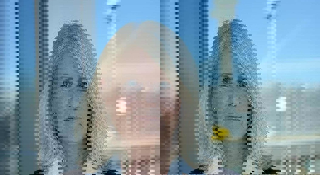 Woman in a blazer with a white shirt stands in front of a glass window overlooking an airport terminal and control tower in the background.