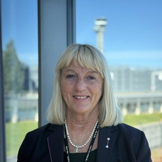 Kvinne med blondt hår i en mørk blazer smiler foran et vindu med utsikt over et åpent landskap og en uklar struktur i bakgrunnen.