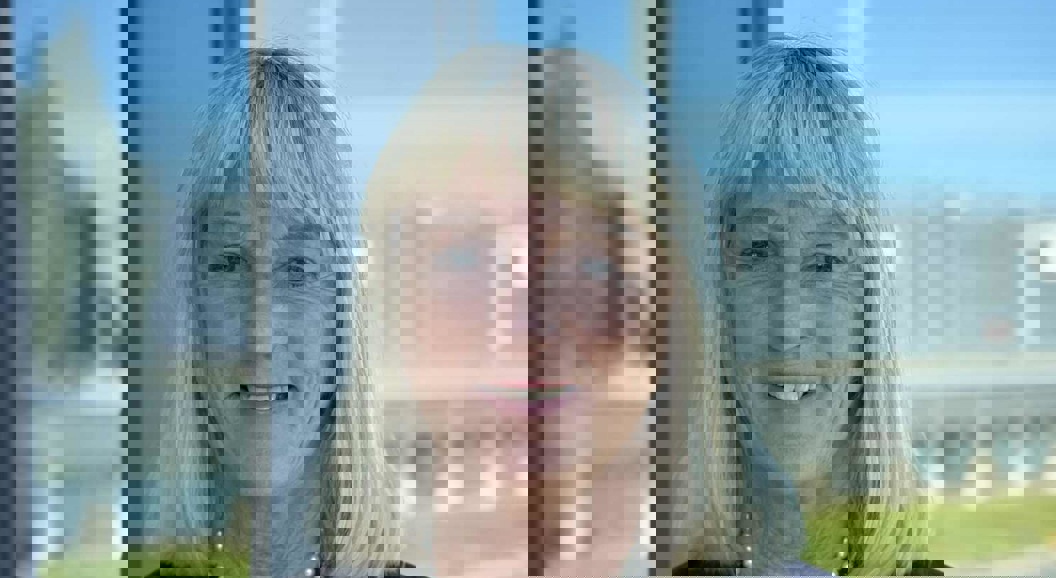 Woman with blonde hair in a dark blazer smiling in front of a window overlooking an open landscape and a blurred structure in the background.