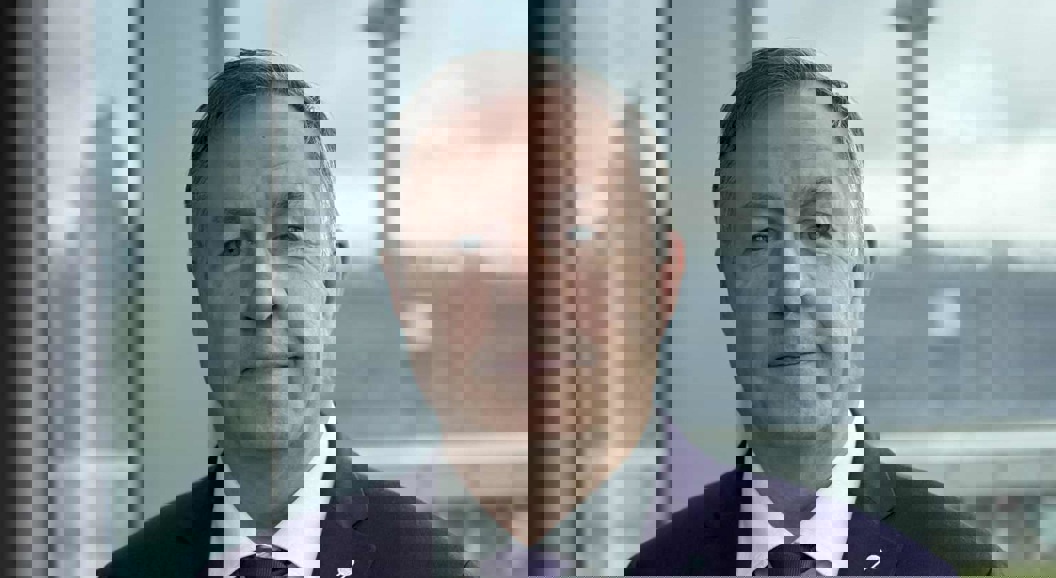 A man in a suit stands in front of a large window overlooking an airport terminal and control tower in the background.