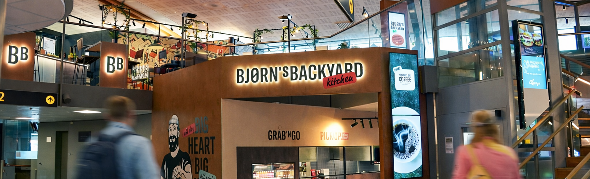 Bjørns Bakgårdskjøkken restaurantinngang i et kjøpesenter, med et veggmaleri av en skjeggete mann som holder mat, med slagordene "Stor Hjerte Stor Meny" og "Grab 'N Go, Pick Up