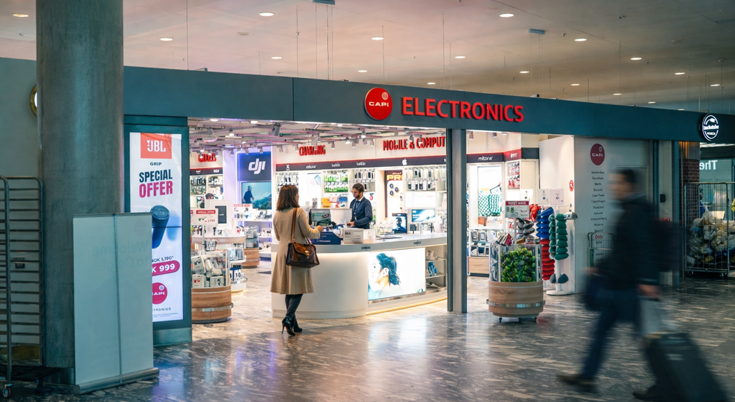 Store employee assisting a customer at a Capi electronics retail counter, surrounded by tech gadgets and souvenirs