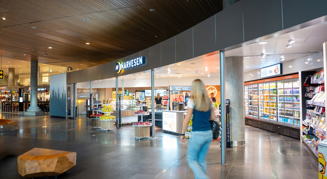 Narvesen kiosk i moderne flyplassinnredning med butikkhyller, kjøledisker og en person som går forbi
