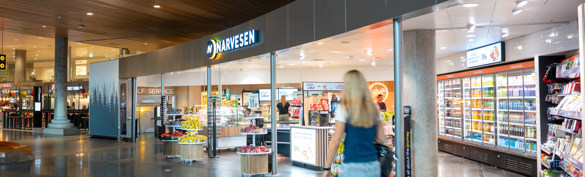 Narvesen kiosk i moderne flyplassinnredning med butikkhyller, kjøledisker og en person som går forbi