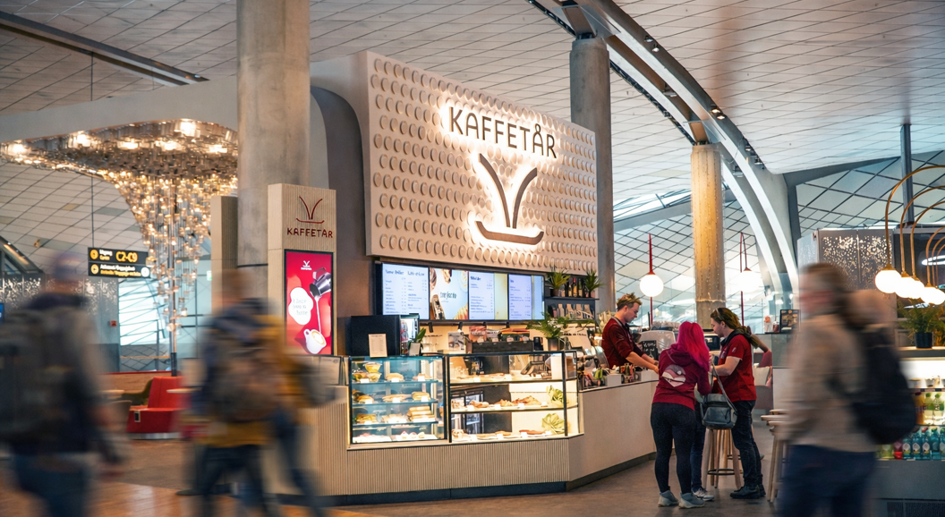 Norsk flyplasslounge med sittegruppe, dekorativ veggdesign med 'Norge', retningsskilt for porter og toaletter, og passasjerer som jobber på bærbare datamaskiner