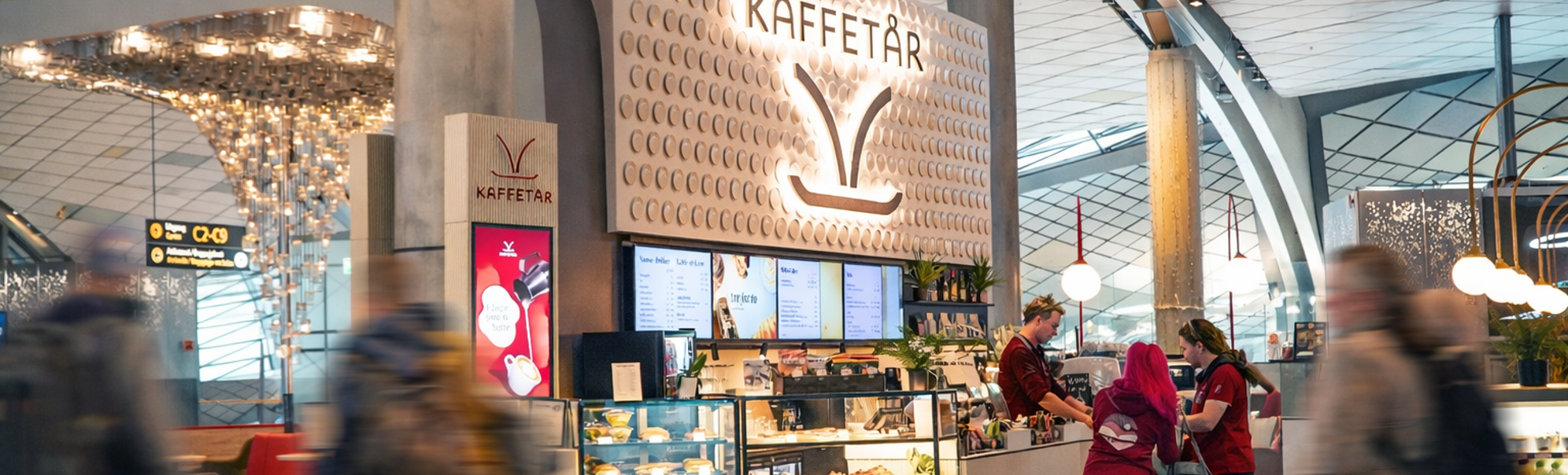 Norsk flyplasslounge med sittegruppe, dekorativ veggdesign med 'Norge', retningsskilt for porter og toaletter, og passasjerer som jobber på bærbare datamaskiner