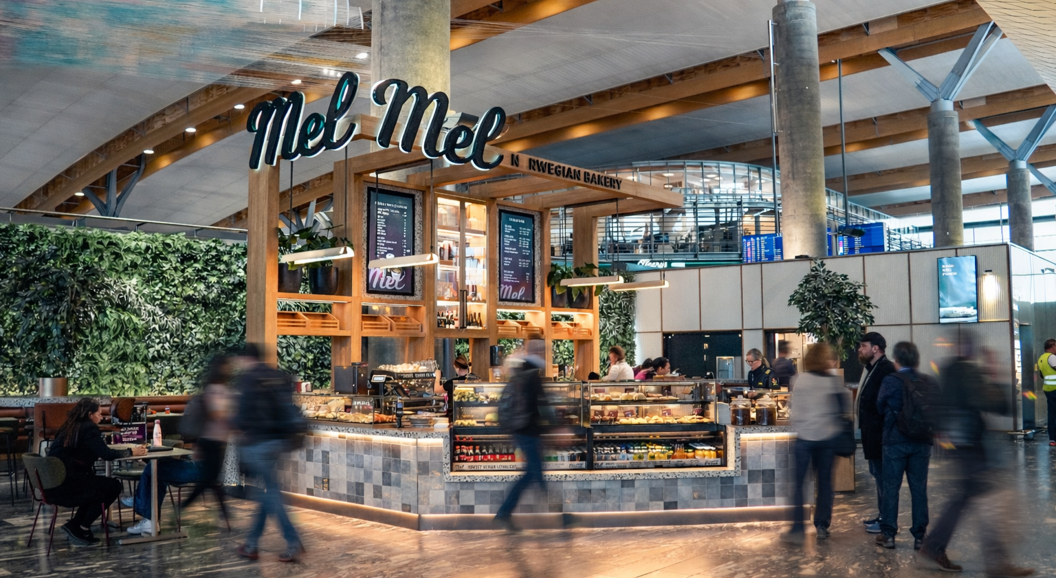 Moderne bakeri og kafé med utstilling av ferske bakverk, kunder som bestiller ved disken, et vennlig skilt "Mel," og en barista som tilbereder kaffe. Lys, moderne interiør med en livlig atmosfære