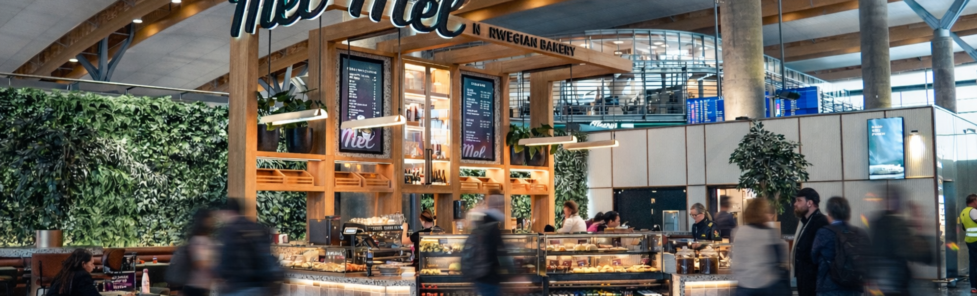Moderne bakeri og kafé med utstilling av ferske bakverk, kunder som bestiller ved disken, et vennlig skilt "Mel," og en barista som tilbereder kaffe. Lys, moderne interiør med en livlig atmosfære