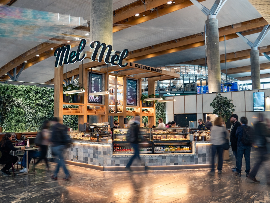 Moderne bakeri og kafé med utstilling av ferske bakverk, kunder som bestiller ved disken, et vennlig skilt "Mel," og en barista som tilbereder kaffe. Lys, moderne interiør med en livlig atmosfære