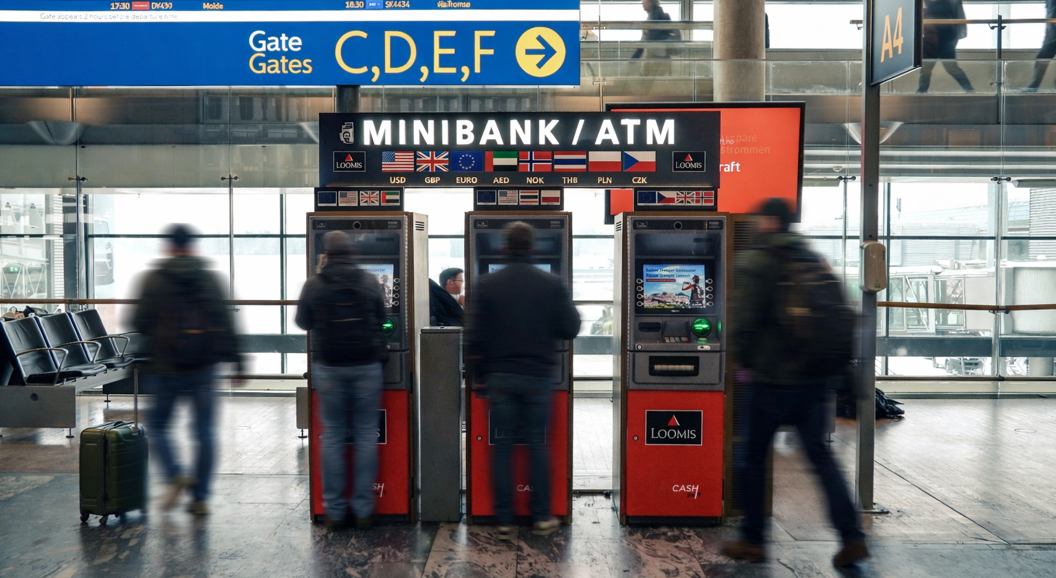 Person using an international ATM at an airport, accessing various global currencies with a digital exchange rate board in the background