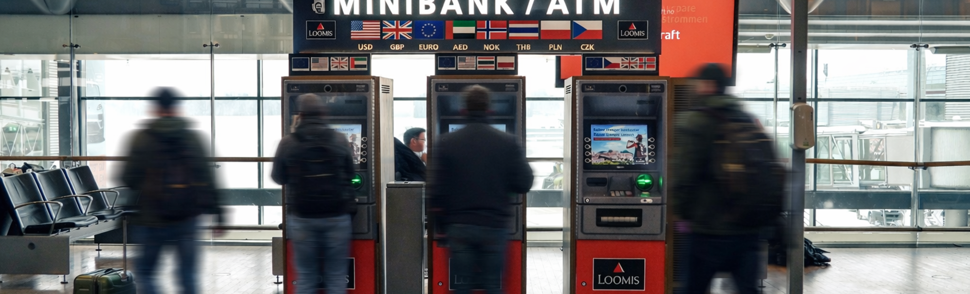 En person som bruker en minibank for utenlandsk valuta på en flyplass, med flere valutaalternativer vist på en skjerm i nærheten, som indikerer internasjonal reise og finansielle transaksjoner