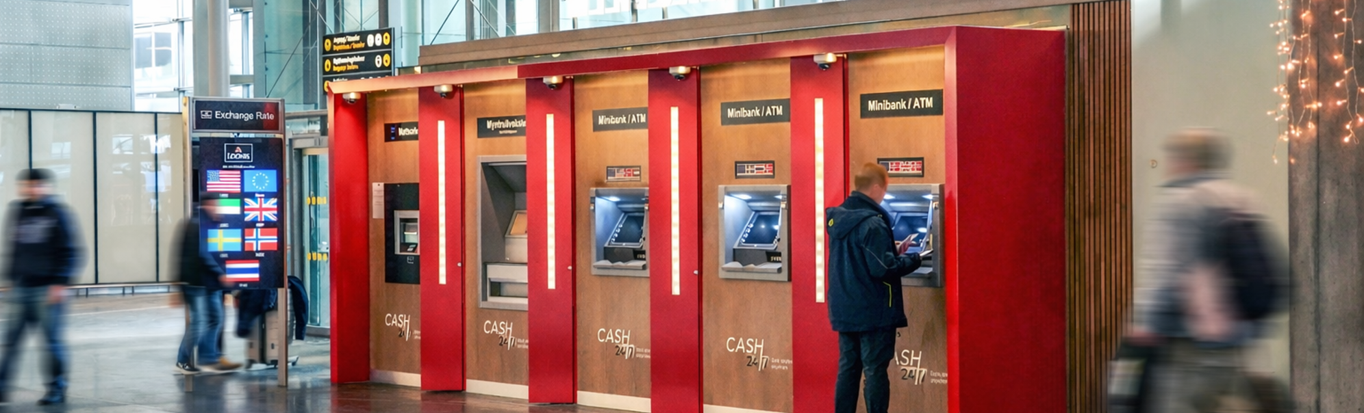 A person using an ATM at an airport, featuring various currency options and an exchange rate display