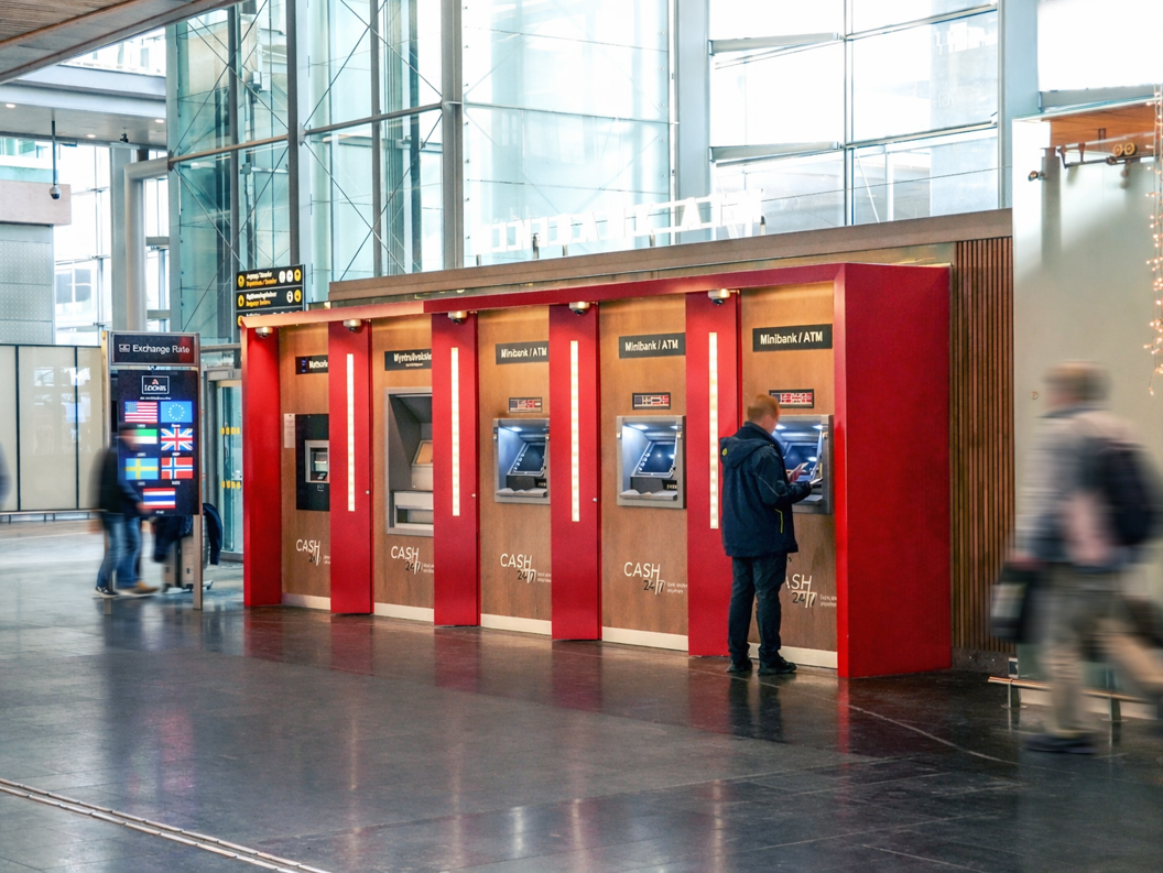 A person using an ATM at an airport, featuring various currency options and an exchange rate display