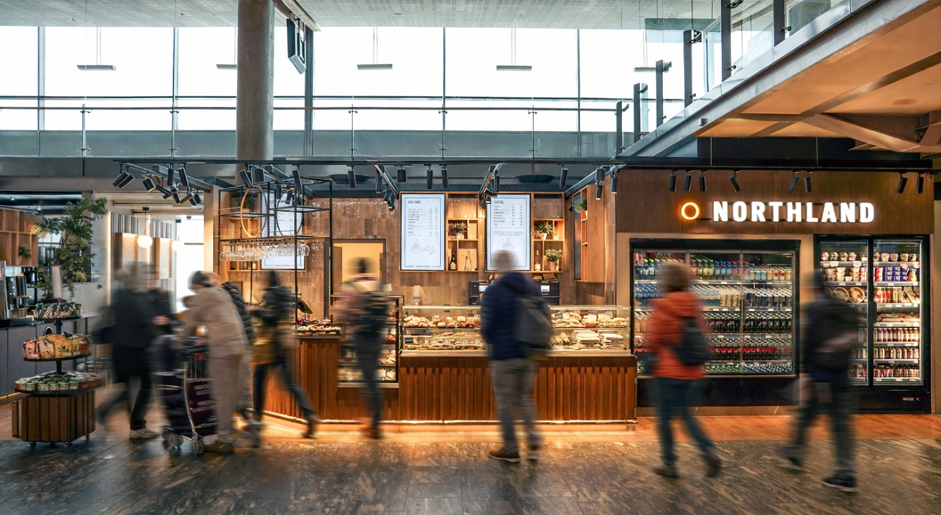 Moderne flyplasskiosk med kunder som ser på drikkevarer og snacks på Northland, med lyst interiør og et utvalg av mat og drikkevarer utstilt