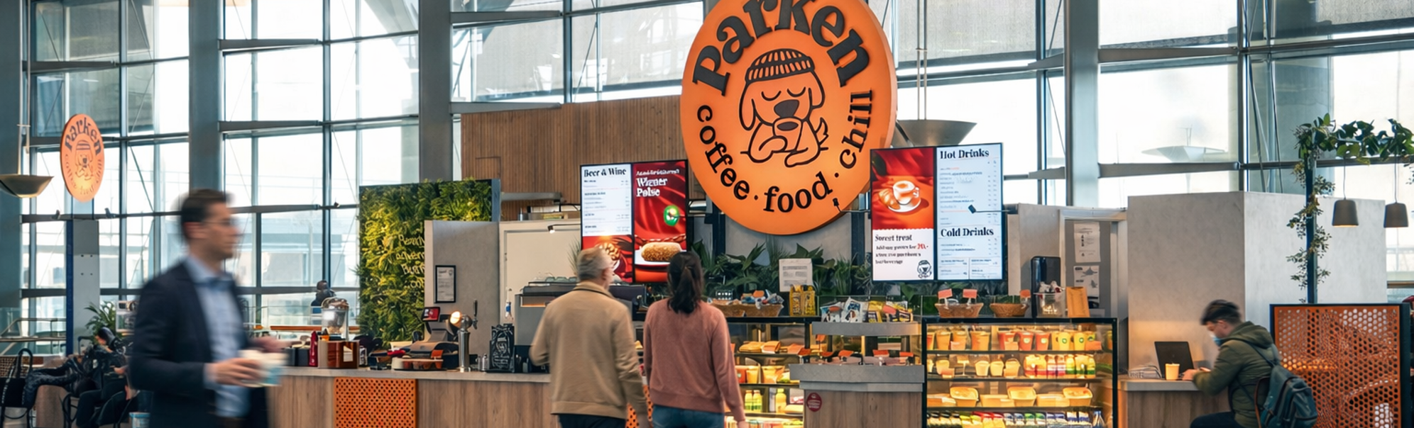 Moderne flyplasskafé med store glassvinduer, med oransje skilt og rikelig med sitteplasser, tilbyr et utvalg av mat og drikke