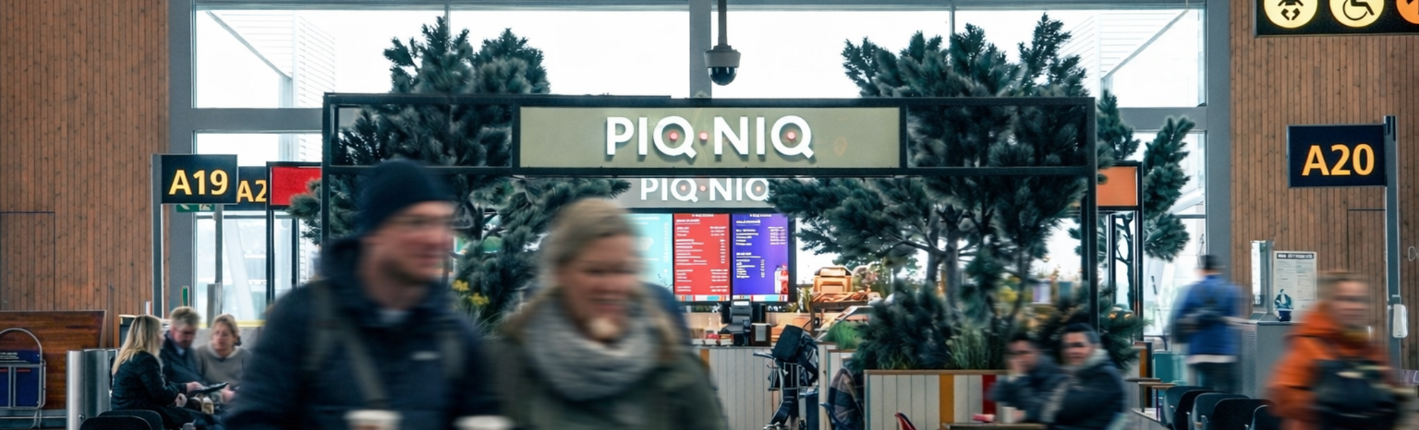 Fargerikt moderne kaféinteriør med skiltet "PIQNIO" og livlige krakker, et nærbilde av en kaffekvern og kopper, ferske bakverk og sandwicher, og dekorative planter som forsterker den innbydende atmosfæren