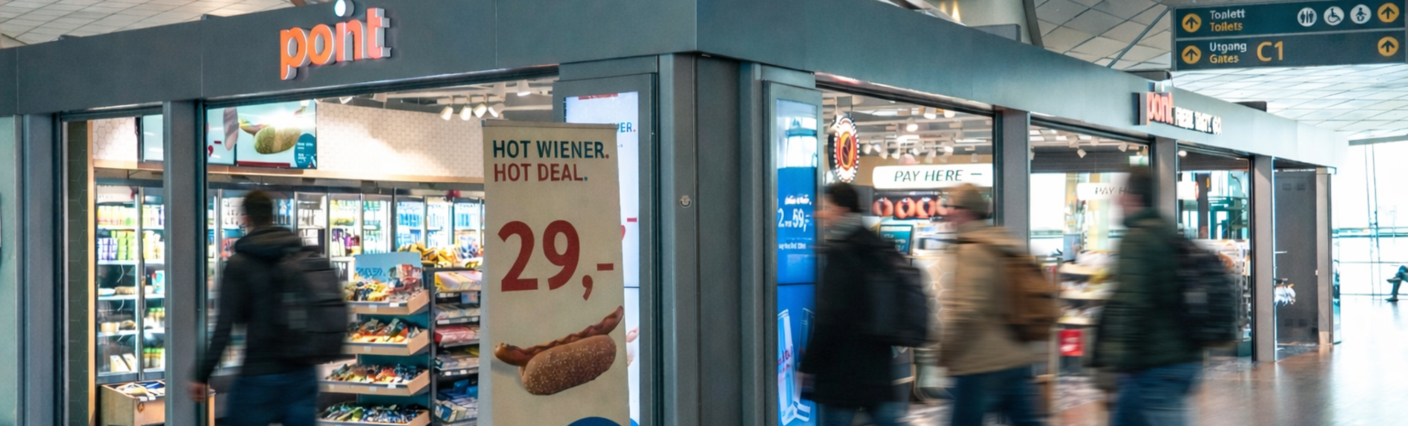 Smilende kassamedarbeider i blå uniform ved en kasse i en nærbutikk, med et reklameskilt i bakgrunnen