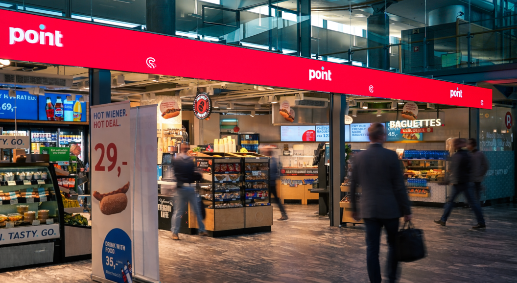 Store sign in an airport with the text "point" and "FRESH. TASTY. GO." displayed above a shop offering magazines and books, with visible ceiling lighting and signage for traveler gates