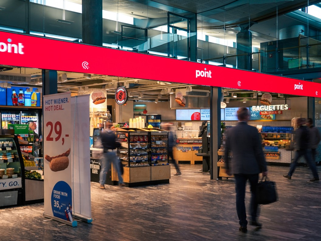 Store sign in an airport with the text "point" and "FRESH. TASTY. GO." displayed above a shop offering magazines and books, with visible ceiling lighting and signage for traveler gates