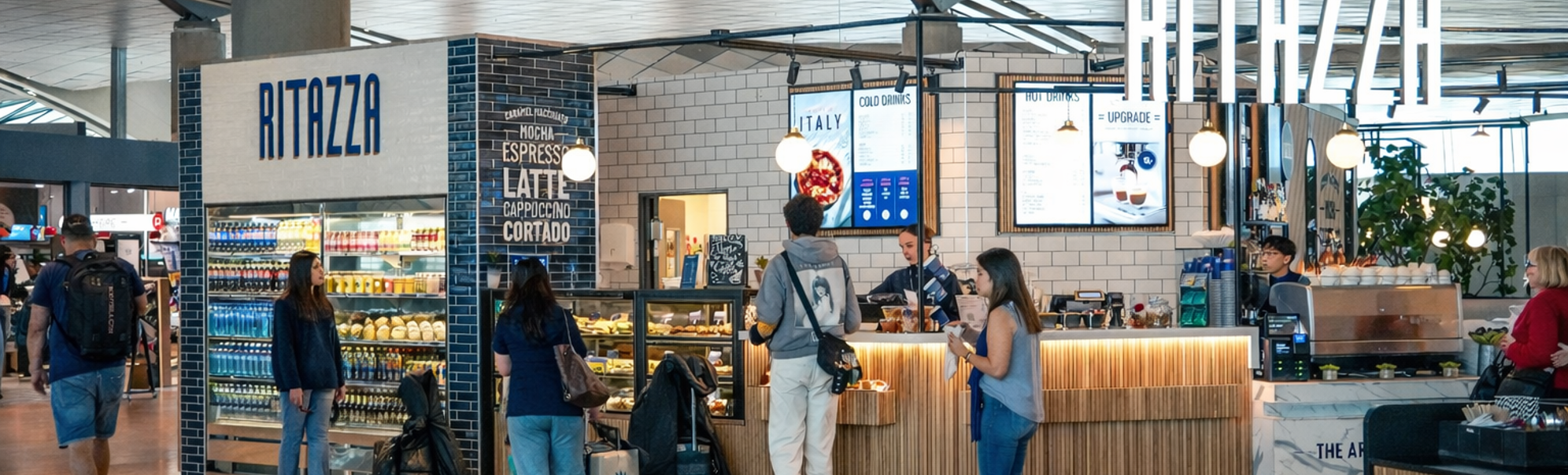 Blå Ritazza kaféskilt omgitt av grønt, med en refleksjon av et moderne interiør i glassvinduet, som antyder en koselig og innbydende atmosfære