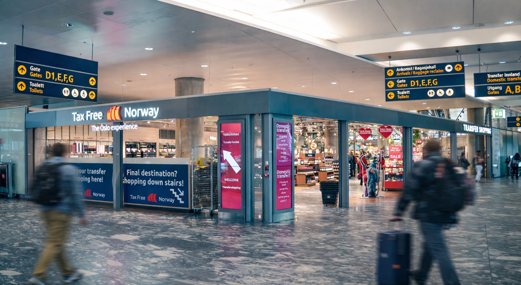 Airport duty-free shop and transfer area at Oslo Airport with travelers and directional signs to gates and toilets