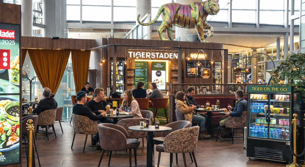Moderne flyplassbar og loungeområde med sitteplasser, med en stor dekorativ tigerskulptur over "Tigerstaden Vinbar" skilt, med gjester og personale synlige, og et retningsskilt for porter E, F, G i bakgrunnen