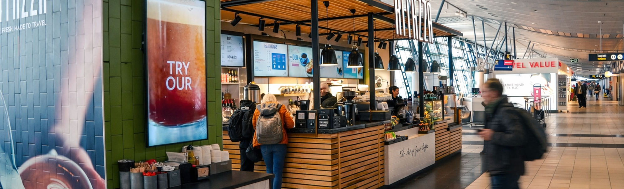 Flyplassterminalens kaffe- og bakverkiosk med reisende som går forbi under moderne buede taklys